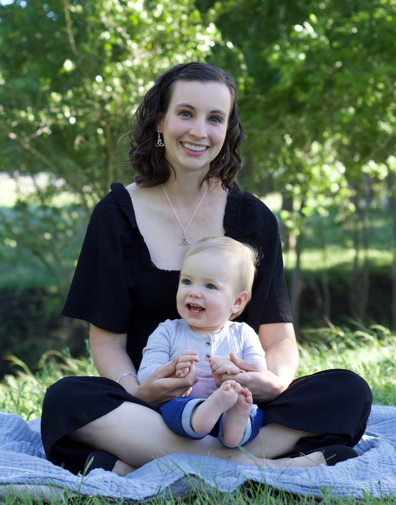 May include: A woman wearing a black top and black pants is sitting on a blue blanket with a baby in her lap. The baby is wearing a light blue and gray shirt and jeans. The woman and baby are smiling at the camera.