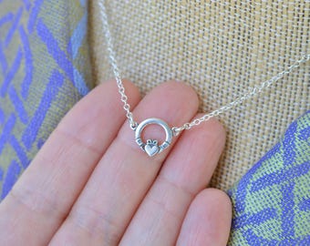 Pode incluir: Um colar Claddagh prateado. O pendente apresenta um coração, mãos e uma coroa, simbolizando amor, lealdade e amizade. O colar é exibido sobre um fundo texturizado com um tecido estampado.