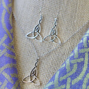 May include: A silver necklace and earrings set featuring a Celtic knot design. The necklace and earrings are on a light brown fabric background.