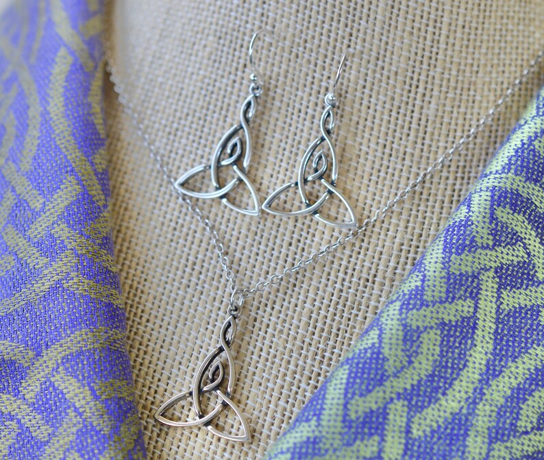 May include: A silver necklace and earrings set featuring a Celtic knot design. The necklace and earrings are on a light brown fabric background.