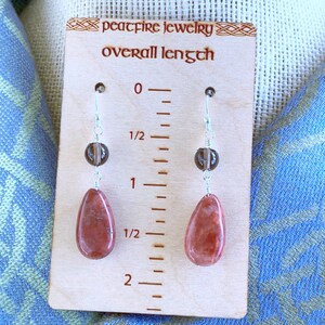 May include: A pair of dangle earrings with a red gemstone and a brown bead. The earrings are approximately 2 inches long. "peacfire jewelry overall length" is written on the wooden background.
