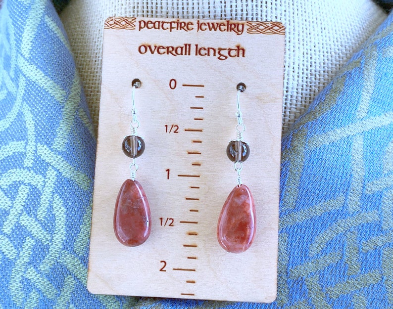 May include: A pair of dangle earrings with a red gemstone and a brown bead. The earrings are approximately 2 inches long. "peacfire jewelry overall length" is written on the wooden background.