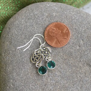 Puede incluir: Un par de pendientes de plata con nudo celta y detalles de cristal verde. Los pendientes se muestran sobre una superficie de piedra gris junto a un centavo estadounidense para la escala.