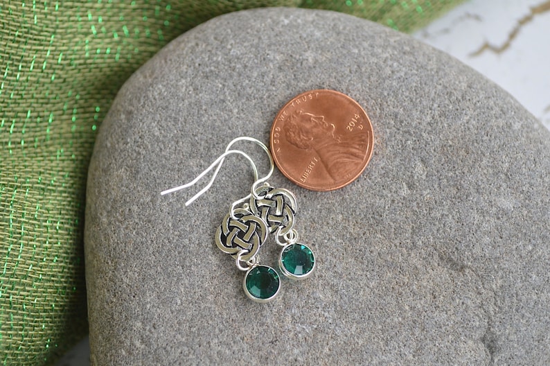 Puede incluir: Un par de pendientes de plata con nudo celta y detalles de cristal verde. Los pendientes se muestran sobre una superficie de piedra gris junto a un centavo estadounidense para la escala.