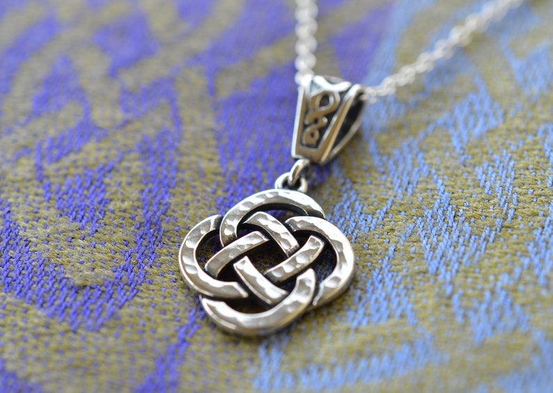 May include: Silver Celtic knot pendant necklace with a hammered texture. The pendant is in the shape of a square knot with four loops. The necklace is on a silver chain.