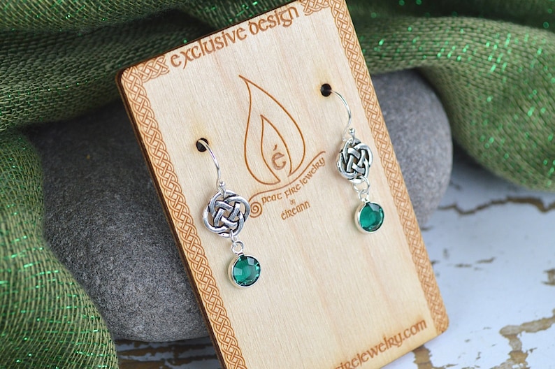 Puede incluir: Pendientes de nudo celta de plata con gotas de cristal verde. Los pendientes se muestran en una tarjeta de madera con el texto "exclusive design" y "peat fire jewelry" en un borde de nudo celta.