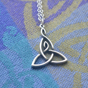May include: A silver Celtic knot pendant necklace with a triangular shape. The pendant is on a blue and green patterned fabric background.