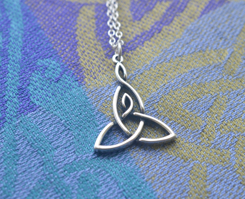 May include: A silver Celtic knot pendant necklace with a triangular shape. The pendant is on a blue and green patterned fabric background.