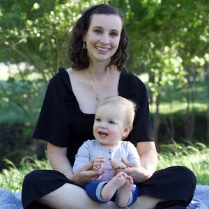 May include: A woman wearing a black top and black pants is sitting on a blue blanket with a baby in her lap. The baby is wearing a light blue and gray shirt and jeans. The woman and baby are smiling at the camera.