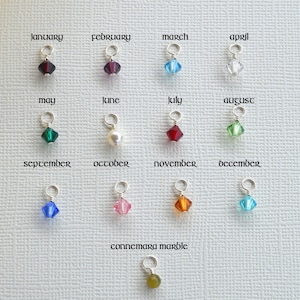 May include: Twelve crystal charms in various colors, each representing a different month of the year. The charms are displayed on a white surface with the month names written above each charm. The charms are small and delicate, and they would make a beautiful addition to any jewelry piece.