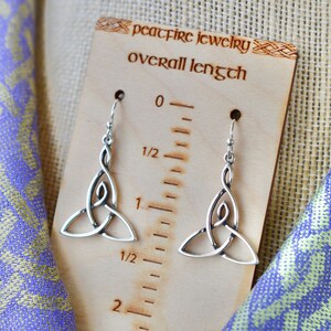 May include: Silver Celtic knot earrings with a drop design. The earrings are shown on a wooden ruler with the text "peacefire jewelry overall length" and measure 1 1/2 inches long.