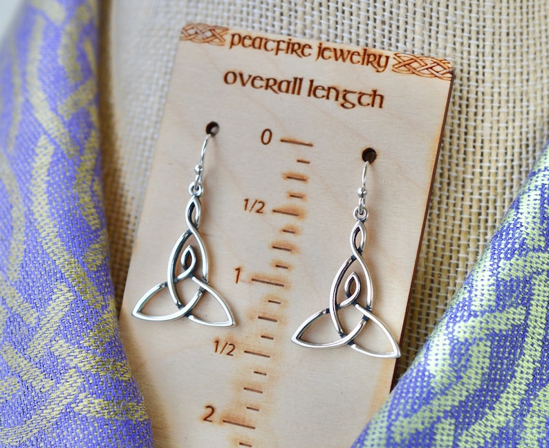 May include: Silver Celtic knot earrings with a drop design. The earrings are shown on a wooden ruler with the text "peacefire jewelry overall length" and measure 1 1/2 inches long.