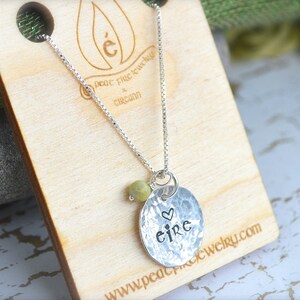 May include: Silver necklace with a hammered disc pendant that says "eire" with a small heart above the word. The necklace is on a wooden surface with a green fabric in the background.