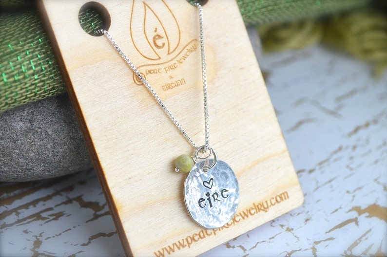May include: Silver necklace with a hammered disc pendant that says "eire" with a small heart above the word. The necklace is on a wooden surface with a green fabric in the background.