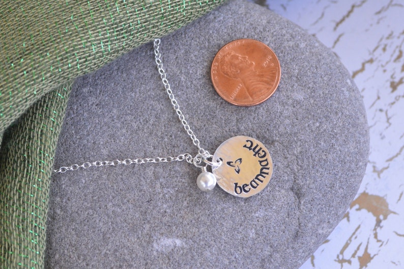 K&ouml;nnte beinhalten: Silberne Halskette mit einem runden Anh&auml;nger, der einen keltischen Knoten und das Wort "Sch&ouml;nheit" in g&auml;lischer Schrift zeigt. Der Anh&auml;nger ist an einer silbernen Kette mit einer kleinen Perle befestigt.