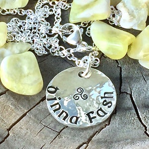 May include: A silver necklace with a round pendant that says "ó  b  inna  Fash" in a Celtic font. The pendant has a Celtic knot design and is surrounded by small, light yellow stones.