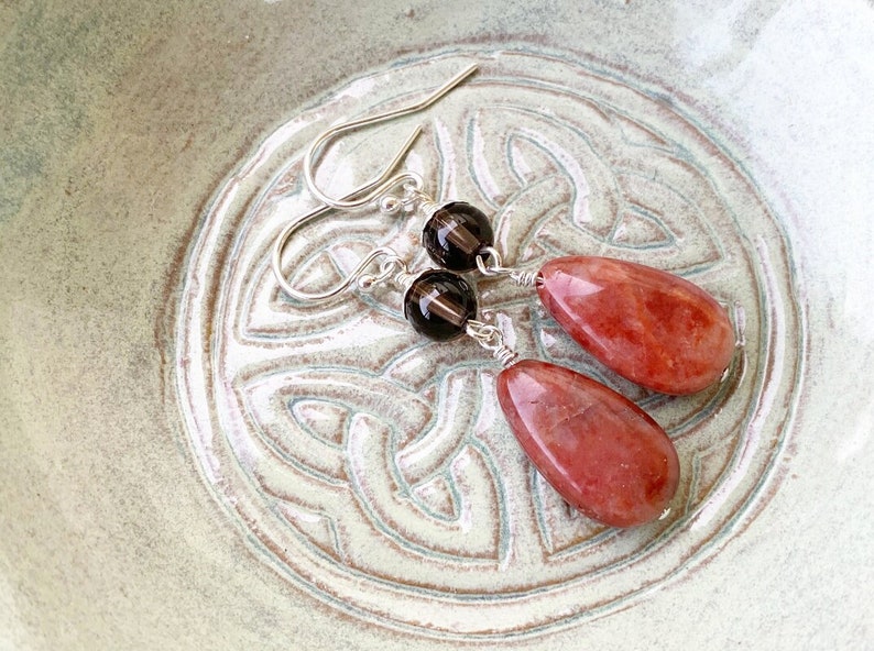 May include: A pair of dangle earrings with a teardrop-shaped red gemstone and a round brown gemstone. The earrings are made with silver wire and have silver hooks.