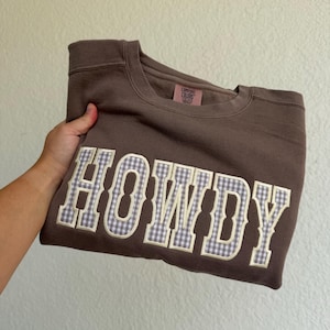 Puede incluir: Una sudadera marrón de cuello redondo con la palabra HOWDY en letras grandes de estilo bloque. Las letras están rellenas con un patrón de cuadros vichy en violeta claro y blanco y delineadas en crema. Una etiqueta de marca Comfort Colors es visible en el cuello.