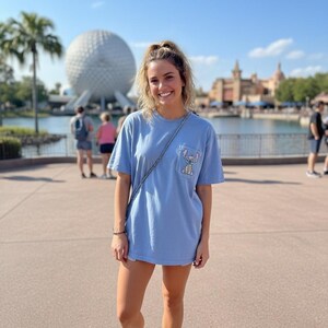 Puede incluir: Una camiseta azul claro con un bolsillo con un gr&aacute;fico de Stitch. La persona lleva zapatillas blancas y un bolso de cadena plateada. El fondo incluye una gran esfera blanca y una masa de agua.