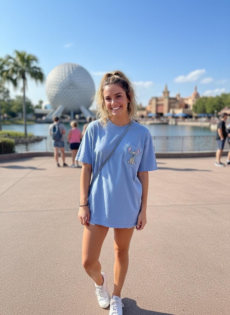 Puede incluir: Una camiseta azul claro con un bolsillo con un gr&aacute;fico de Stitch. La persona lleva zapatillas blancas y un bolso de cadena plateada. El fondo incluye una gran esfera blanca y una masa de agua.