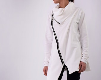 May include: A white long-sleeved shirt with a zipper detail and a cowl neck. The shirt is asymmetrical and has a long, flowing hem. The model is wearing black leggings and black boots.