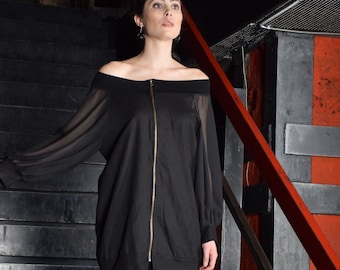 May include: A woman wearing a black off-the-shoulder long-sleeved top with a zip down the front and a black skirt. She is standing on a set of stairs with a red handrail.