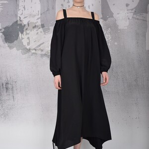 May include: A black off-the-shoulder dress with long sleeves and a square neckline. The dress has a flowing, asymmetrical hemline and is paired with silver sneakers. The background is a textured gray and white wall.