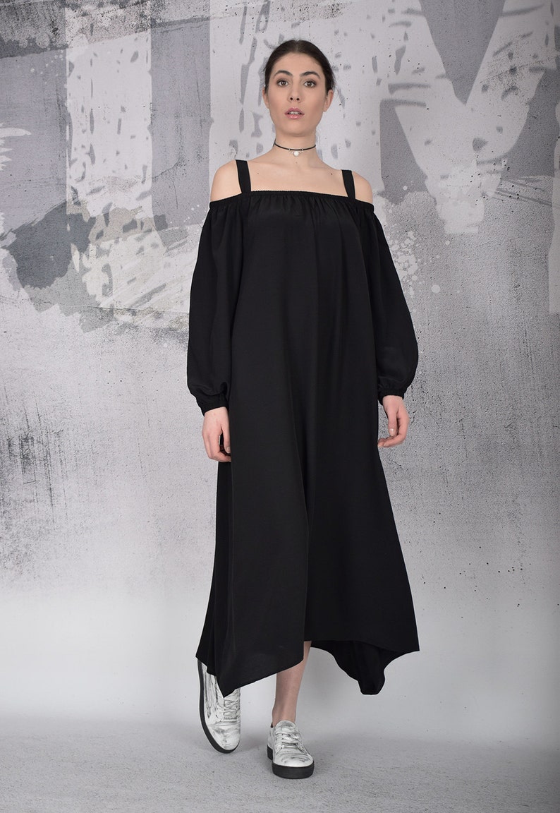 May include: A black off-the-shoulder dress with long sleeves and a square neckline. The dress has a flowing, asymmetrical hemline and is paired with silver sneakers. The background is a textured gray and white wall.