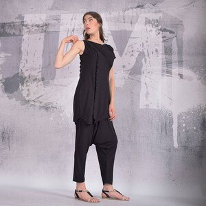 Puede incluir: Una mujer con un top negro sin mangas con un dobladillo con volantes y pantalones negros. Est&aacute; de pie frente a una pared gris con salpicaduras de pintura blanca.