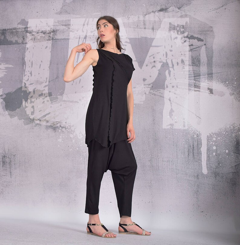 Puede incluir: Una mujer con un top negro sin mangas con un dobladillo con volantes y pantalones negros. Est&aacute; de pie frente a una pared gris con salpicaduras de pintura blanca.
