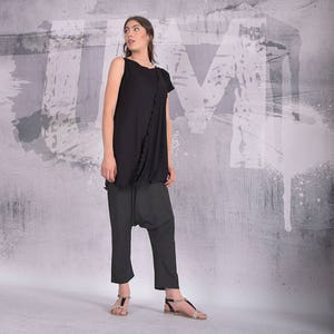 Puede incluir: Una mujer con un top negro sin mangas con un detalle de volantes y pantalones negros. Est&aacute; de pie frente a una pared gris con salpicaduras de pintura blanca.