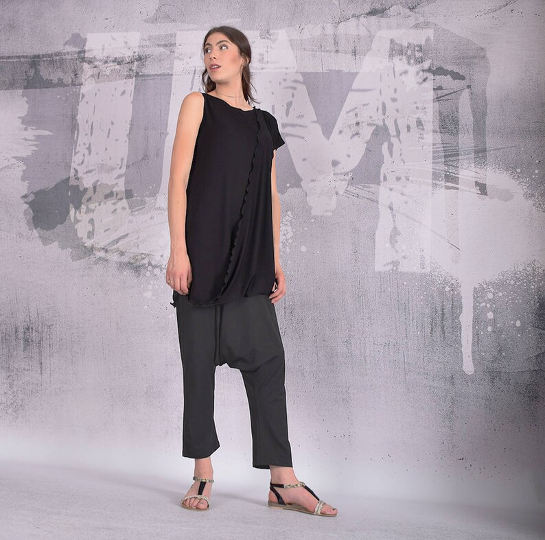 Puede incluir: Una mujer con un top negro sin mangas con un detalle de volantes y pantalones negros. Est&aacute; de pie frente a una pared gris con salpicaduras de pintura blanca.