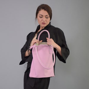 May include: A pink leather tote bag with white stitching and a top handle. The bag is held by a person wearing a black jacket.