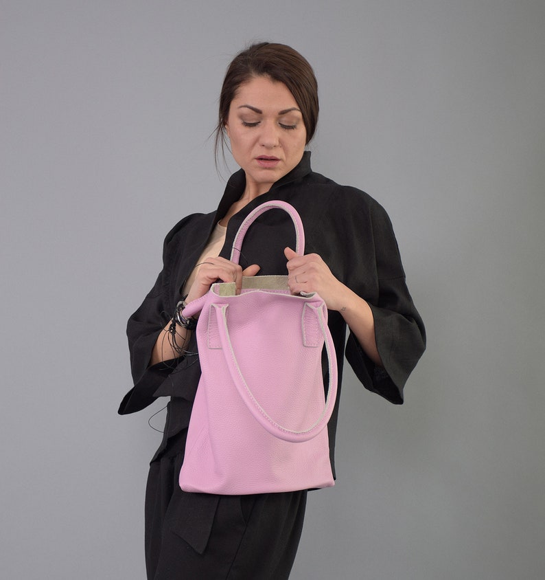 May include: A pink leather tote bag with white stitching and a top handle. The bag is held by a person wearing a black jacket.