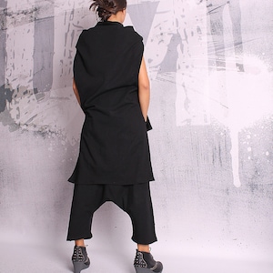 May include: A black sleeveless tunic with a long, flowing hemline and a pair of black pants with a wide leg. The outfit is worn by a person standing in front of a white and gray textured wall.