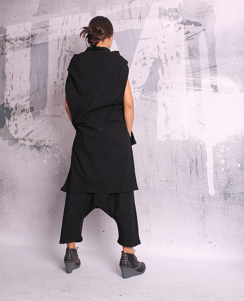 May include: A black sleeveless tunic with a long, flowing hemline and a pair of black pants with a wide leg. The outfit is worn by a person standing in front of a white and gray textured wall.