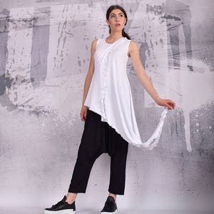 Op de afbeelding: Een wit topje zonder mouwen met een gedrapeerd ontwerp en een zwarte broek met een wijde pasvorm. Het model draagt zwarte sneakers.