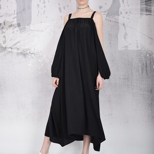 May include: A black, off-the-shoulder dress with wide straps and long sleeves. The dress has a flowing, asymmetrical hemline and is paired with white sneakers. The background is a textured gray and white wall.