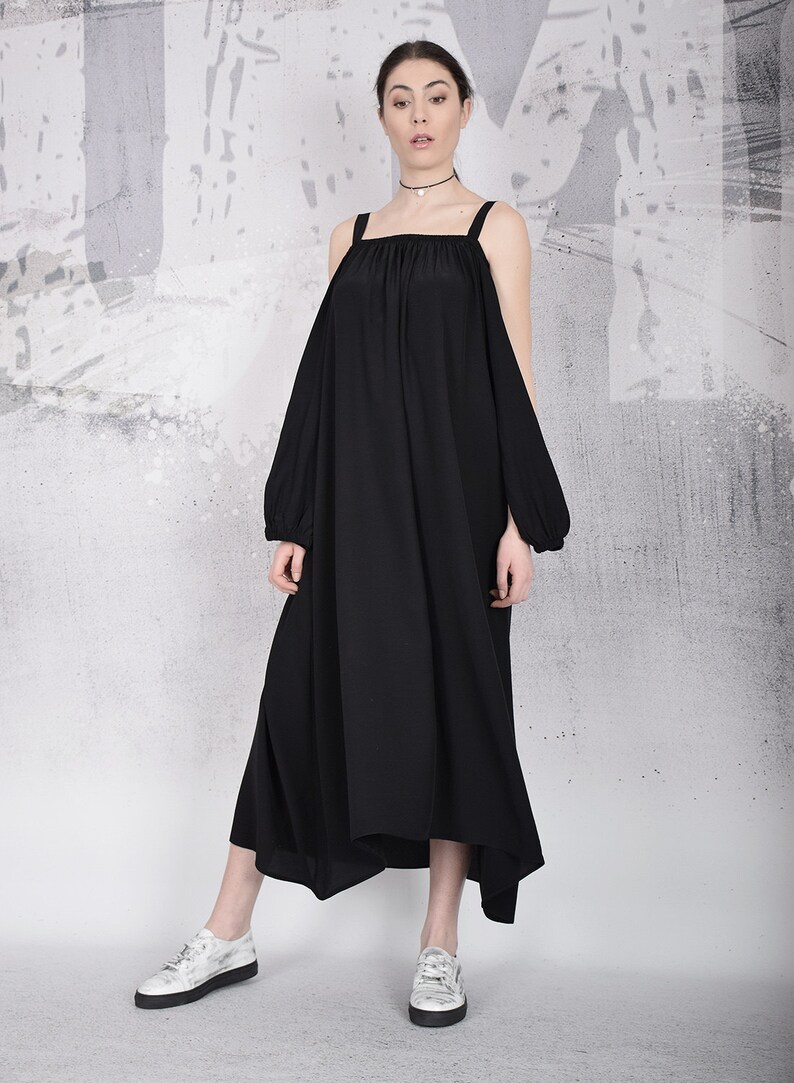 May include: A black, off-the-shoulder dress with wide straps and long sleeves. The dress has a flowing, asymmetrical hemline and is paired with white sneakers. The background is a textured gray and white wall.