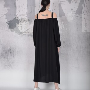 May include: A black off-the-shoulder dress with long sleeves and a flowing skirt. The dress has wide shoulder straps and is paired with white shoes. The background is a textured gray and white wall.
