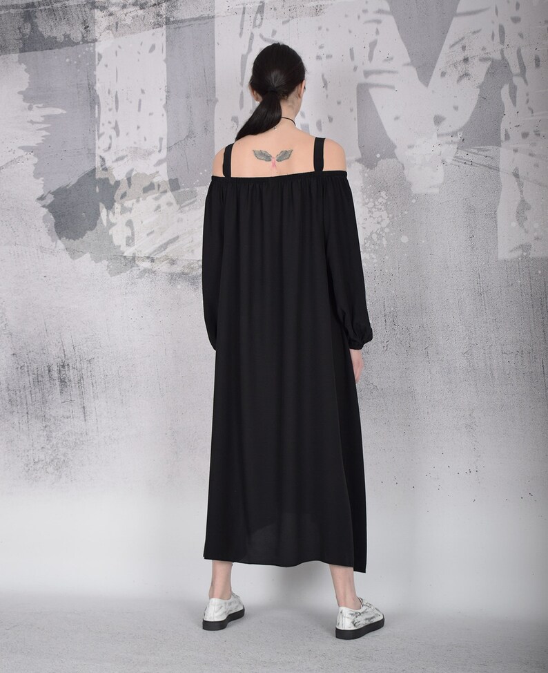 May include: A black off-the-shoulder dress with long sleeves and a flowing skirt. The dress has wide shoulder straps and is paired with white shoes. The background is a textured gray and white wall.
