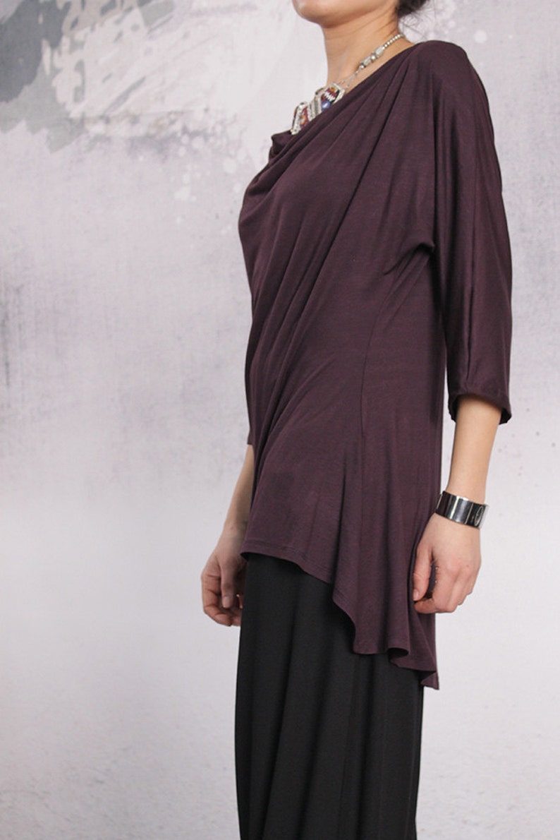 Puede incluir: Una blusa de manga larga, de color morado oscuro, drapeada con cuello de pico y un dobladillo alto-bajo. La blusa est&aacute; hecha de una tela suave y fluida.