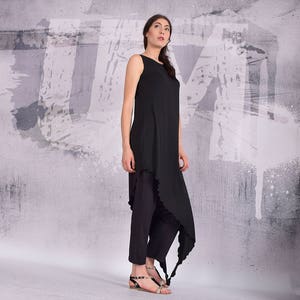 Puede incluir: Una t&uacute;nica negra sin mangas con un dobladillo festoneado y un drapeado largo y fluido. La t&uacute;nica se combina con pantalones negros y sandalias negras.