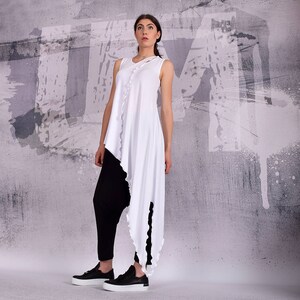 Op de afbeelding: Een wit asymmetrisch tuniek met een gegolfde zoom. De top heeft een ronde hals en is mouwloos. Het model draagt een zwarte broek en zwarte sneakers.