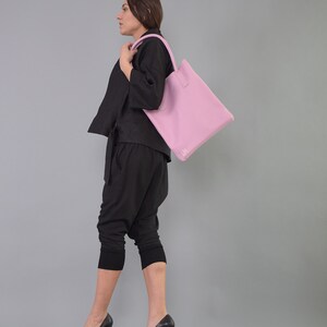 May include: A woman wearing black pants and a black top is carrying a light pink tote bag with a white logo on the front.