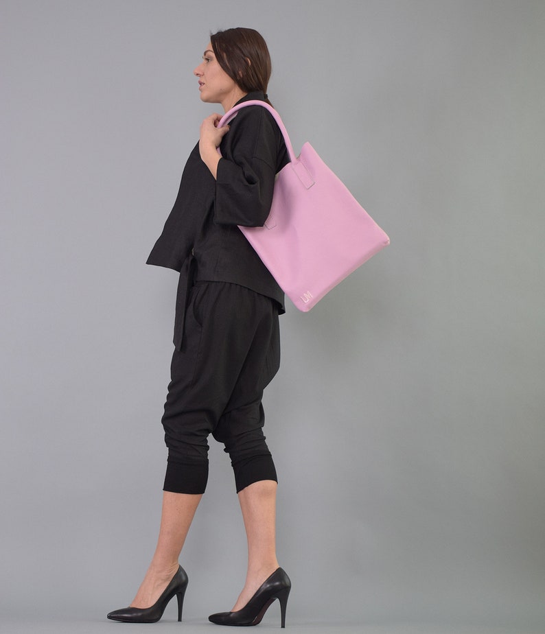 May include: A woman wearing black pants and a black top is carrying a light pink tote bag with a white logo on the front.