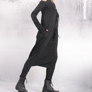 Pode incluir: Um macacão preto de manga comprida com um detalhe de renda na frente e um design de ombro cortado. O macacão tem um caimento solto e é combinado com botas pretas de tornozelo.