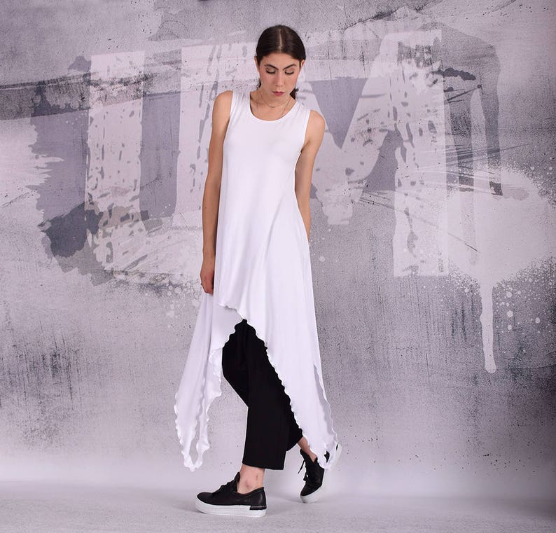 Op de afbeelding: Een witte, mouwloze tuniek met een high-low zoom. De tuniek is gemaakt van een zachte, vloeiende stof en heeft een ontspannen pasvorm. Het model draagt een zwarte broek en zwarte sneakers.