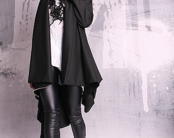 May include: A black asymmetrical jacket with a long, flowing hemline. The jacket is worn over a white shirt and black leggings. The person is wearing black ankle boots.