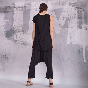 Puede incluir: Un top negro de un solo hombro con un dise&ntilde;o drapeado y pantalones de har&eacute;n negros a juego. La modelo lleva el atuendo sobre un fondo gris con salpicaduras de pintura blanca.